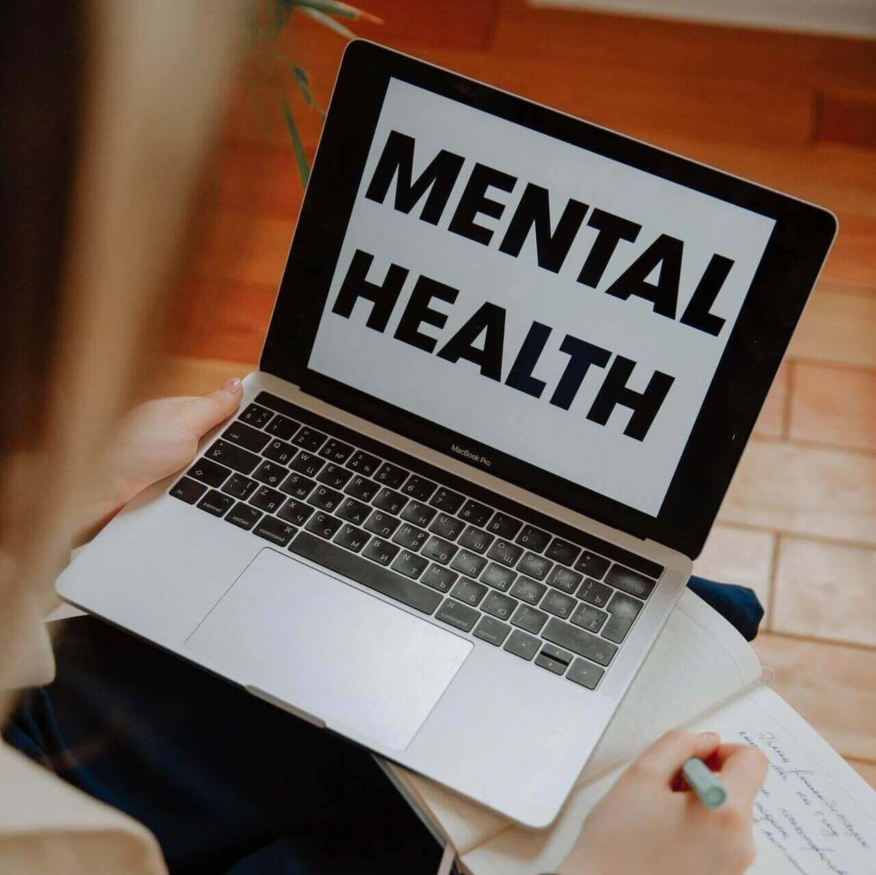 A female psychotherapist working on a laptop displaying the heading 'mental health' and writing down notes in a notebook